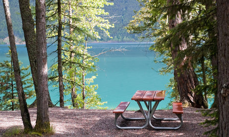 Imagine your RV rental parked nearby! A beautiful picnic table by a clear turquoise lake, framed by lush trees.