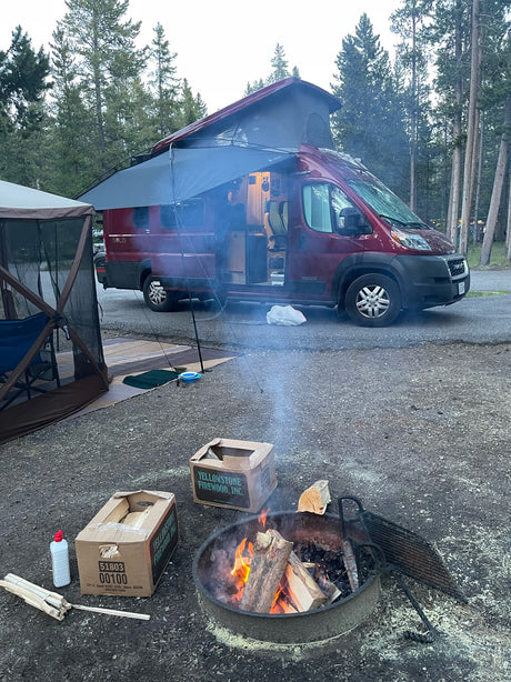 RV rental camping trip: Red camper van with awning, screen tent, and Yellowstone firewood by a crackling campfire.