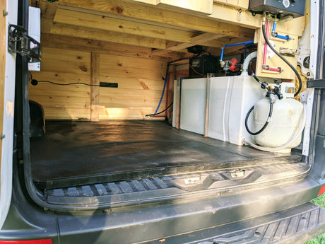 View inside an RV rental van's rear utility area: wood-paneled space with fresh water tank, pump, propane, and water heater.