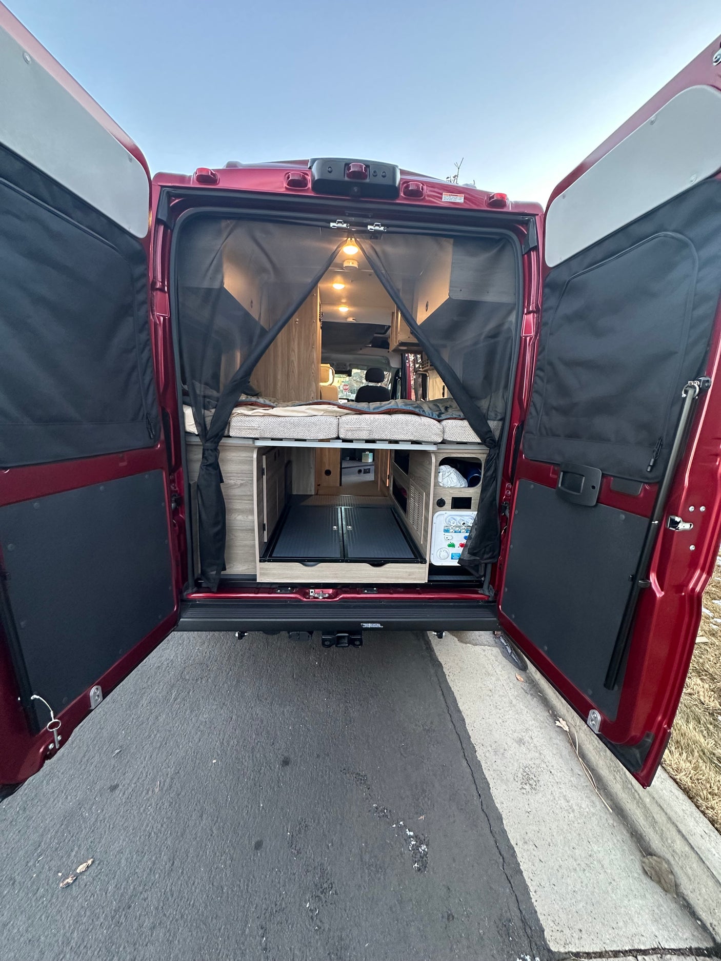 A red RV rental with its back doors open, showcasing a cozy bed, built-in storage, and mesh screen for mobile adventures.