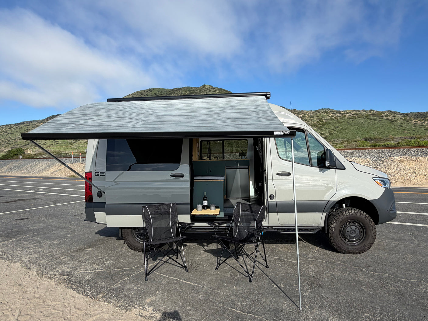 Grey rv rental camper van with extended awning, chairs, and table. Ideal for camping & outdoor adventures by hills.