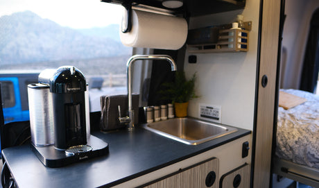 This rv rental's compact kitchen boasts a Nespresso machine, sink, and paper towel dispenser, with a bed visible beyond.