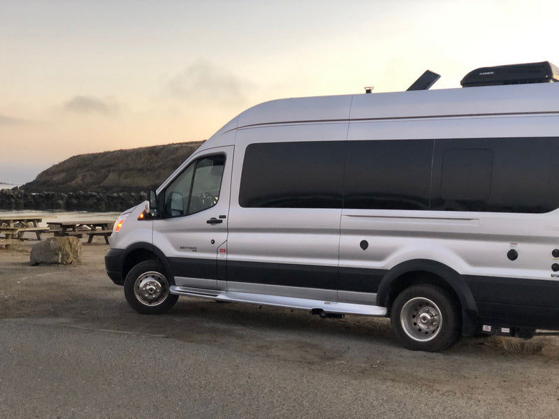  sits by a towering red rock formation where people are climbing.|A man with two French Bulldogs next to a silver Ford Transit RV rental by a lake with bare trees. Great for road trips!|Silver RV rental camper van parked by the ocean at dusk — photo 4