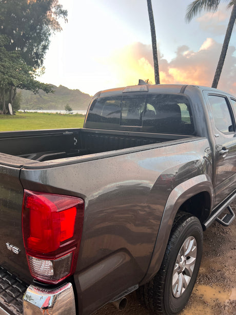 A gray pickup truck, covered in rain droplets, overlooks a serene bay at sunset with palm trees. Perfect for adventure, like an RV rental!