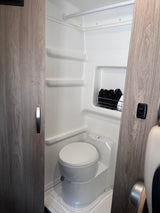 Compact white bathroom inside an RV rental, featuring a toilet, storage shelves, and a shower area.