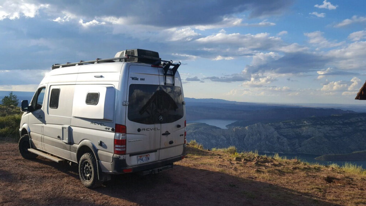 Silver Winnebago Revel rv rental parked on a hill overlooking a vast lake and mountain landscape under cloudy skies.|Explore this RV rental's cozy interior: front seats
