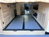 Inside an RV rental: versatile rear storage with textured floor panels and wood-grain cabinetry below the bed.