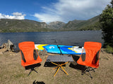 A scenic lake and mountain view features a paddleboard, two orange chairs, and a table. Perfect for an RV rental adventure!