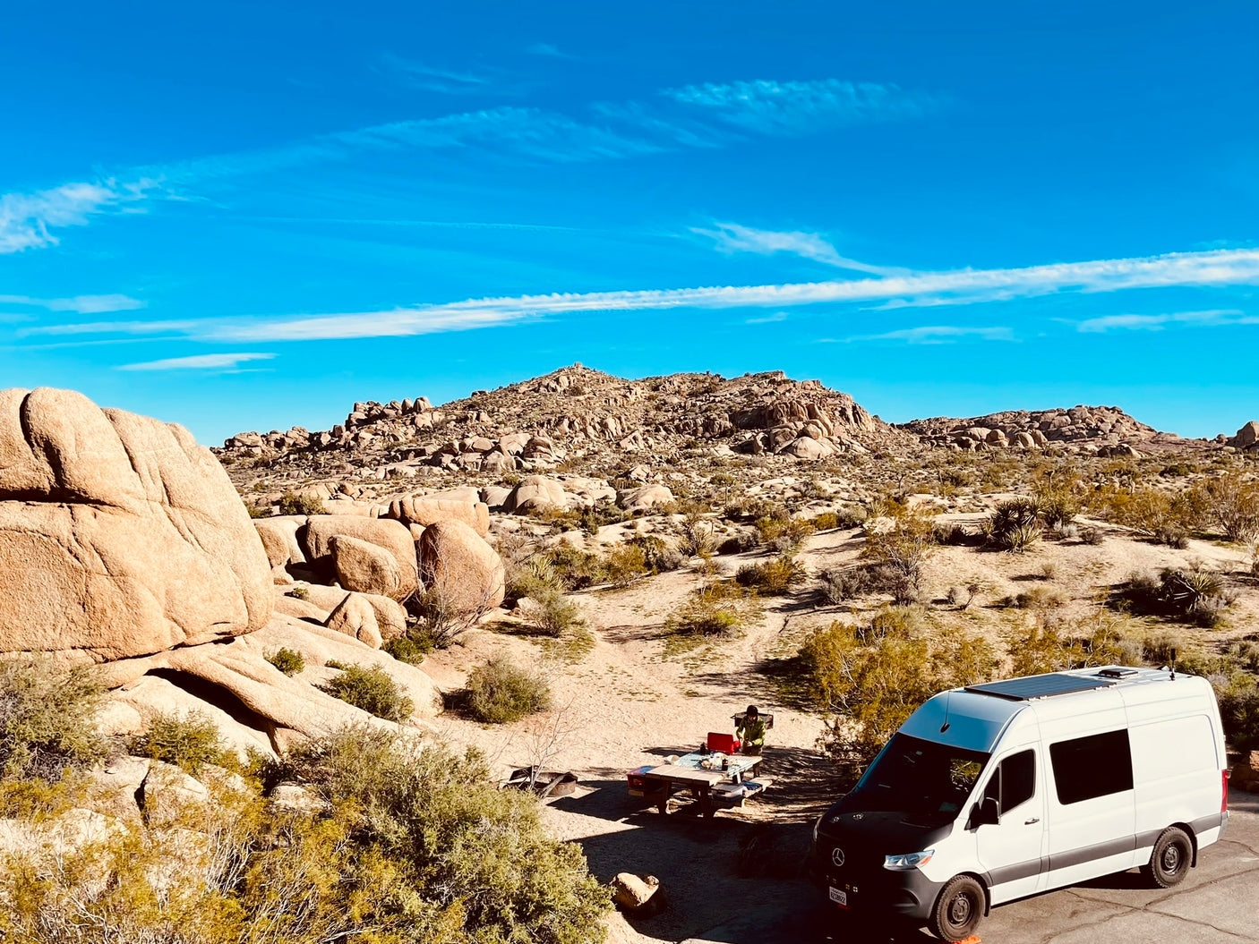 Explore the desert with an RV rental! White camper van at a scenic campsite with rock formations & a person at a picnic table.