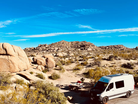 Explore the desert with an RV rental! White camper van at a scenic campsite with rock formations & a person at a picnic table.