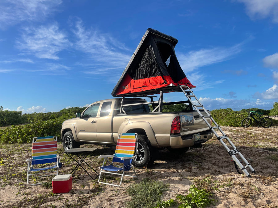 Your next RV rental adventure awaits! Tan Tacoma with rooftop tent, chairs, & cooler set up for camping.