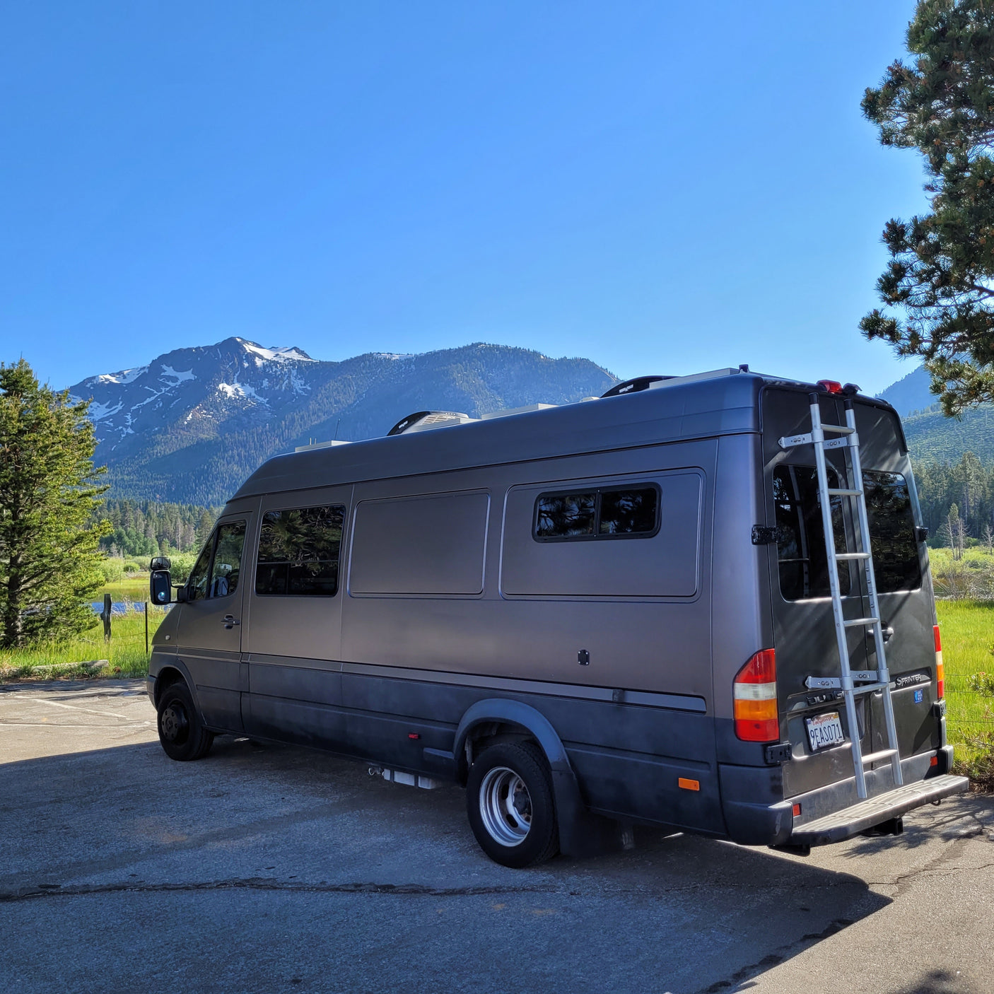 Explore nature! This dark gray RV rental camper van is parked with stunning snow-capped mountains and a lake in the background.