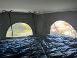 Cozy RV rental pop-top interior: a dark bed with string lights, and arched windows showing lush greenery and houses.