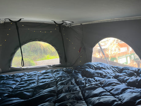 Cozy RV rental pop-top interior: a dark bed with string lights, and arched windows showing lush greenery and houses.