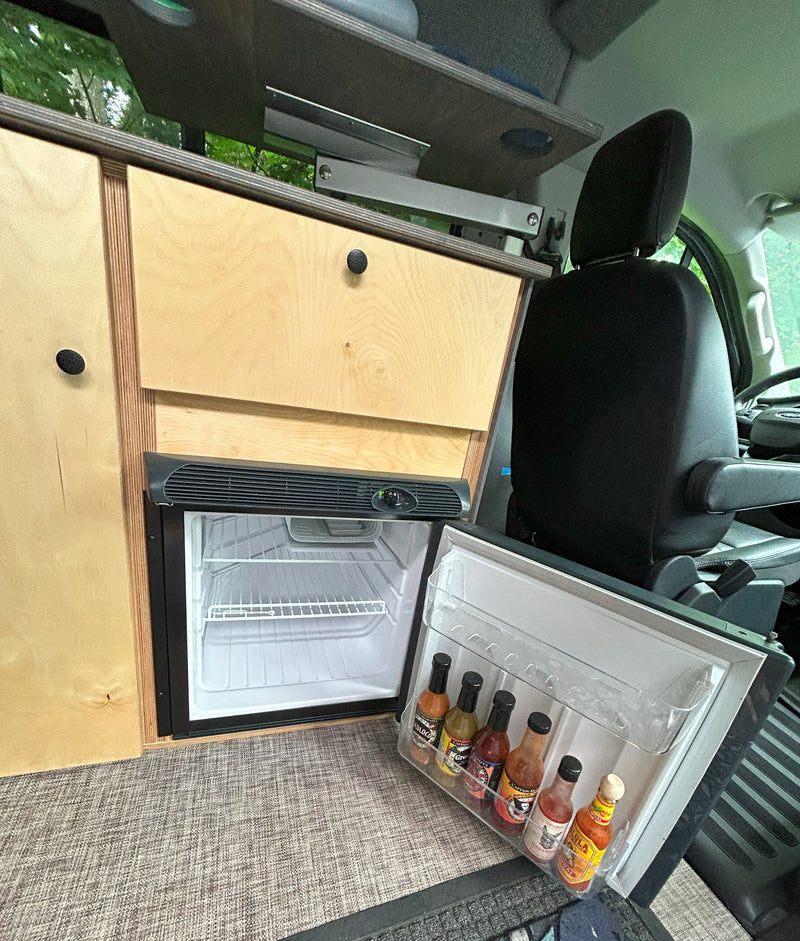 Compact RV rental kitchen with an open fridge showcasing various hot sauces, custom wood cabinetry, and a driver's seat. — photo 10