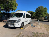 White Mercedes rv rental van parked at a sunny campsite, leveled and hooked to utilities, with other RVs nearby.