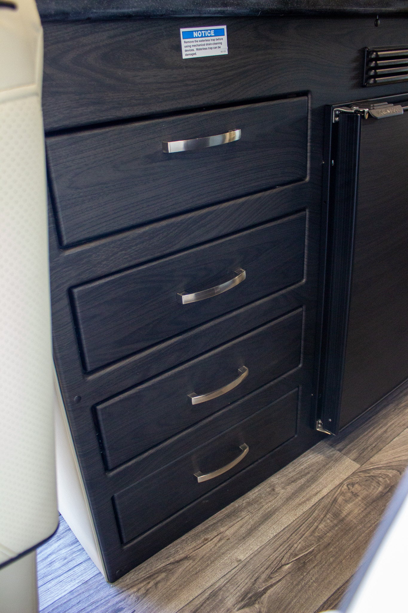 Interior view of an RV rental, showing dark cabinetry with four drawers, a compact fridge, and a waterless trap notice.