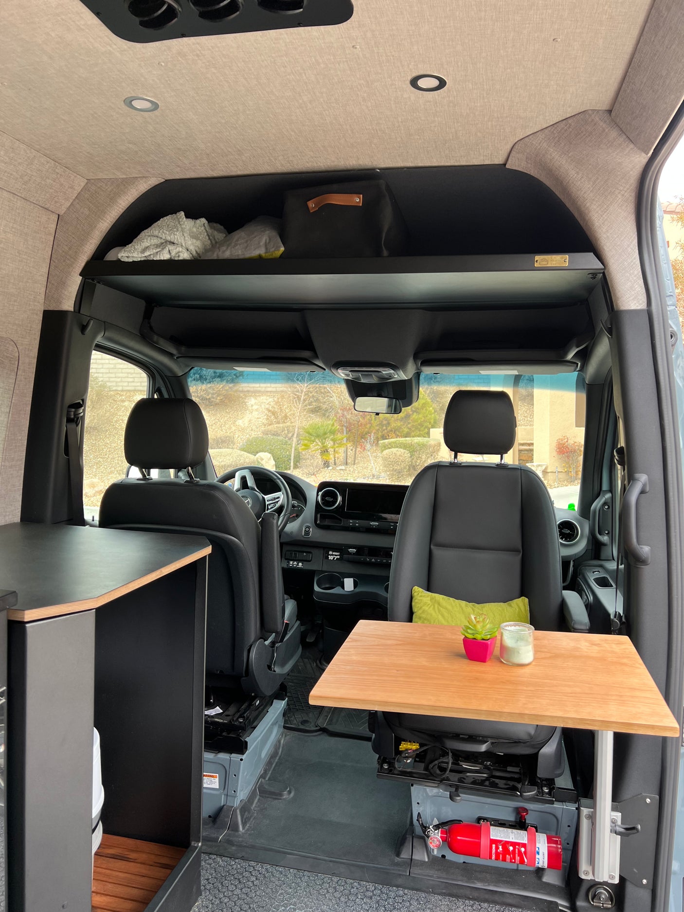 This RV rental camper van interior has an overhead shelf, wood table, and safety features like a fire extinguisher.
