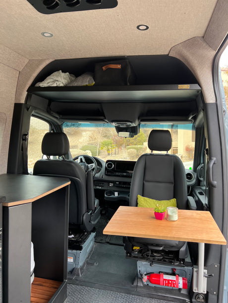 This RV rental camper van interior has an overhead shelf, wood table, and safety features like a fire extinguisher.