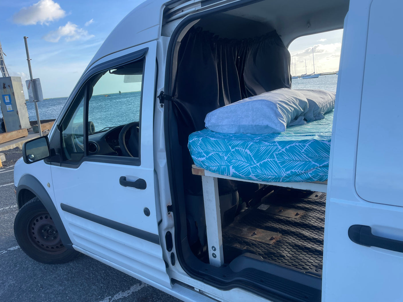 A white camper van with an open side door, showcasing a cozy bed inside with an ocean view. Perfect RV rental for travel.