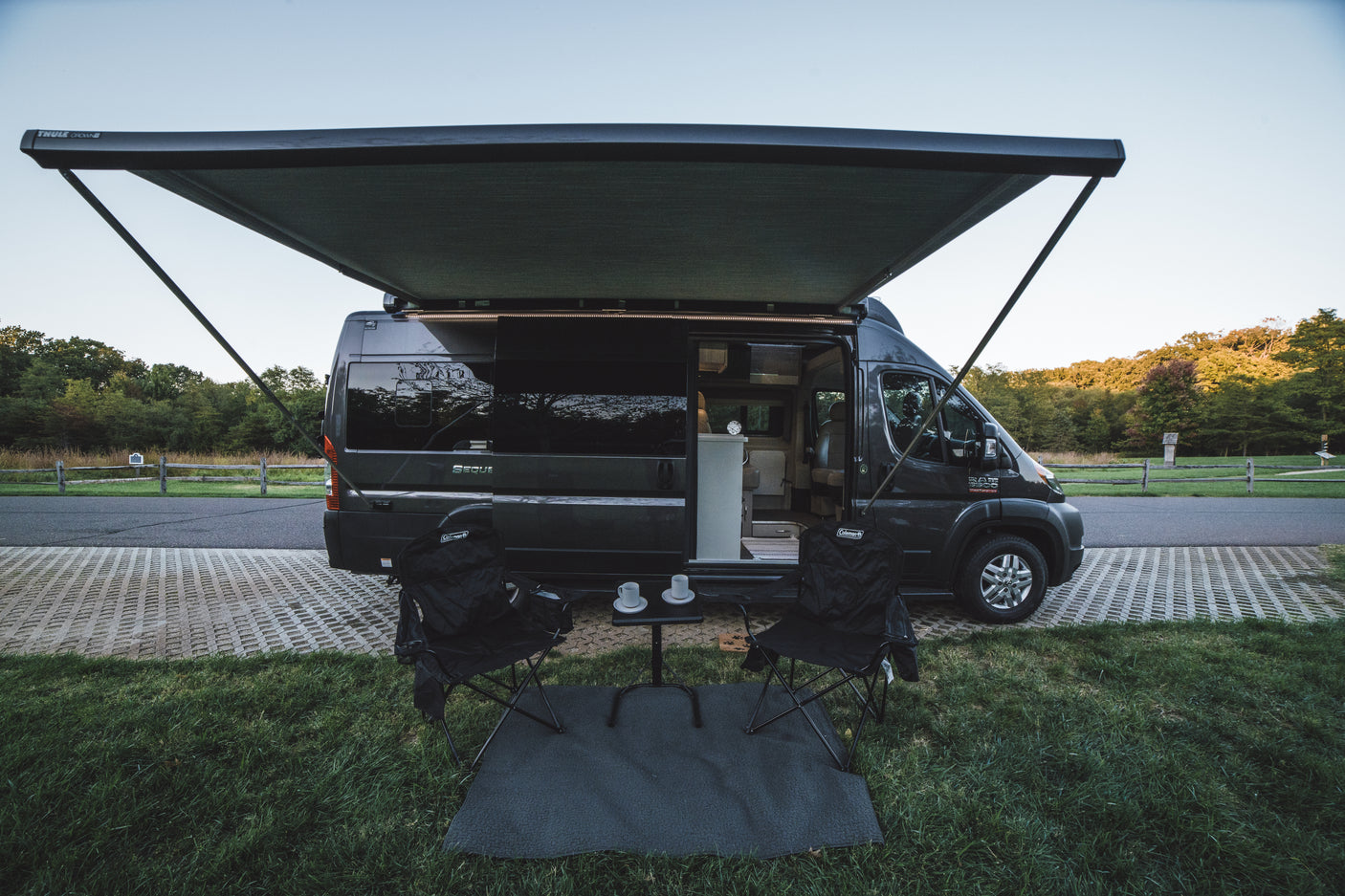 Scenic RV rental setup: A gray camper van with an extended awning, chairs, and table on green grass, ready for camping.
