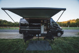 Scenic RV rental setup: A gray camper van with an extended awning, chairs, and table on green grass, ready for camping.