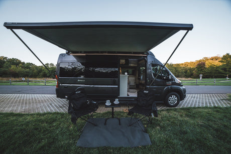 Scenic RV rental setup: A gray camper van with an extended awning, chairs, and table on green grass, ready for camping.