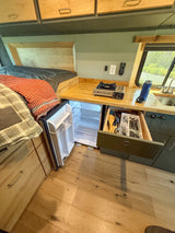 Inside an RV rental: a compact kitchen with a portable stove, sink, open fridge, and cozy bed, ideal for van life.