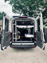 Customized camper van with open rear doors, revealing a versatile interior featuring a suspended bed, storage, and essentials for RV rental.