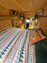 Cozy RV rental interior with a patterned bed, warm wood paneling, and a compact kitchen area with sink and storage.