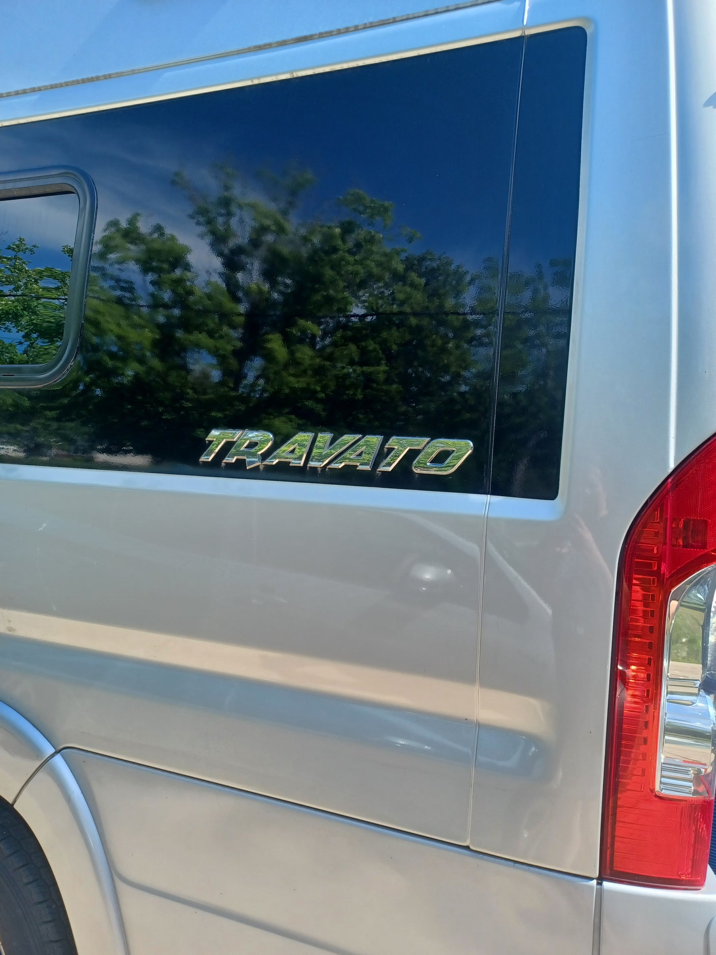 A white Travato RV side with a dark window reflecting trees and the "TRAVATO" emblem. Red taillight visible. Ideal for rv rental.