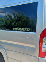 A white Travato RV side with a dark window reflecting trees and the "TRAVATO" emblem. Red taillight visible. Ideal for rv rental.