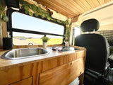 Inside a cozy RV rental: two sinks, wooden counter, and a scenic view outside a large window. Perfect for van life adventures.