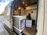 Custom RV rental interior featuring a modern kitchen, open mini-fridge, and cozy seating. Wood ceiling, white walls.