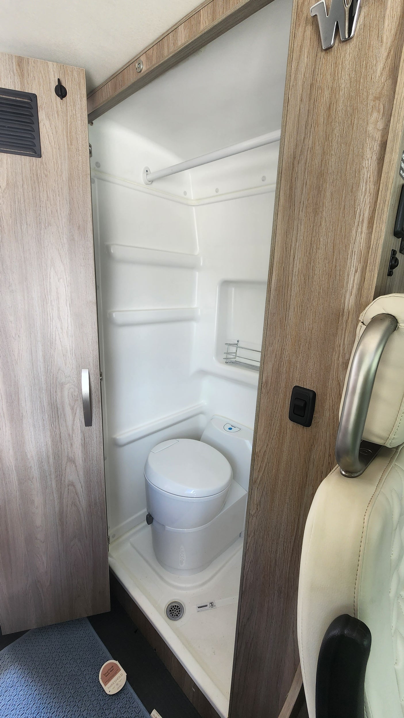 A compact, all-white wet bath in an RV rental, showcasing a toilet, shower pan with a visible drain, and a small wall-mounted rack.