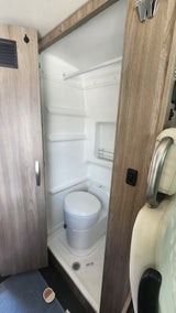 A compact, all-white wet bath in an RV rental, showcasing a toilet, shower pan with a visible drain, and a small wall-mounted rack.