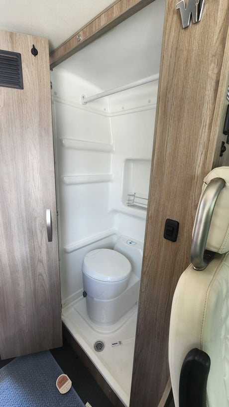 A compact, all-white wet bath in an RV rental, showcasing a toilet, shower pan with a visible drain, and a small wall-mounted rack.
