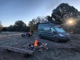 This dark RV rental campervan is parked by a warm campfire and chairs, inviting adventure under a clear sky.