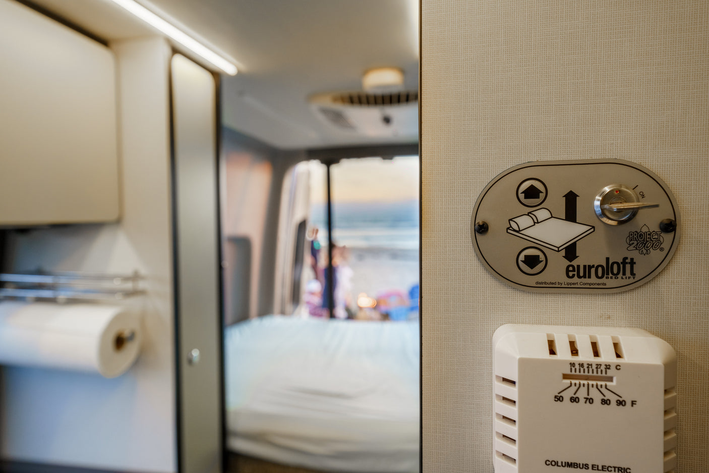 This RV rental interior features a Euroloft bed lift control panel, thermostat, and a serene beach scene visible through the rear.