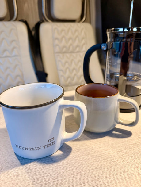Two mugs ("On Mountain Time") and a French press coffee maker inside an RV rental. Cozy camper interior for your road trip.
