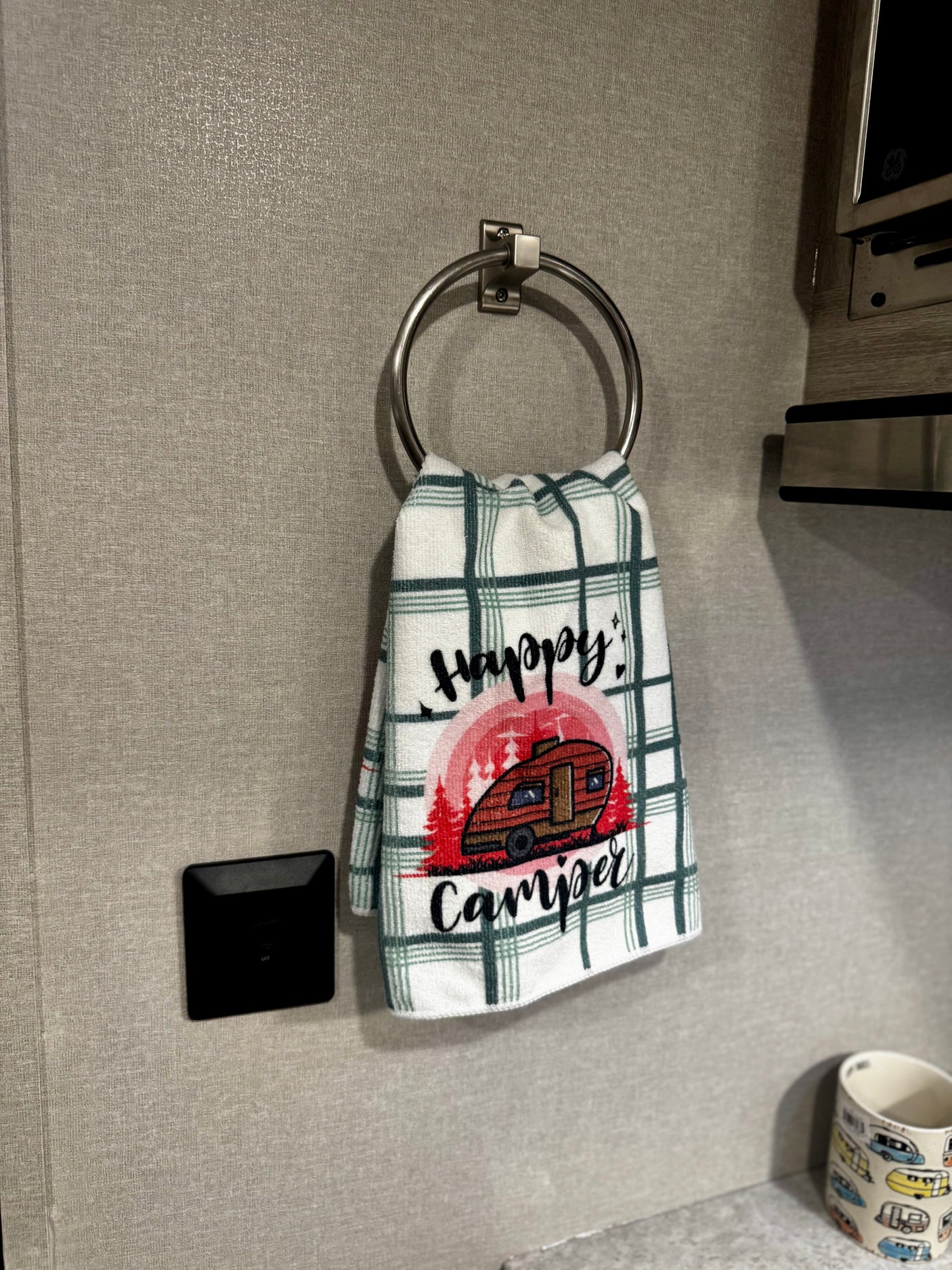 Inside an RV rental: A plaid "Happy Camper" towel hangs by a control panel and countertop mug.