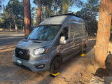 Grey Ford Transit RV rental, leveled on yellow blocks at a sunny forest campsite. Trees surround the vehicle.