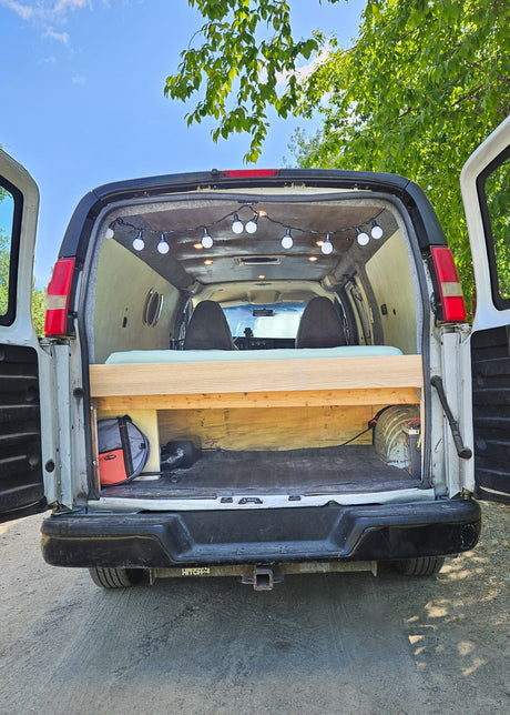 Explore with this custom camper van conversion, ready for adventure! Rear doors open, showing the cozy bed, string lights, and storage in this RV rental.