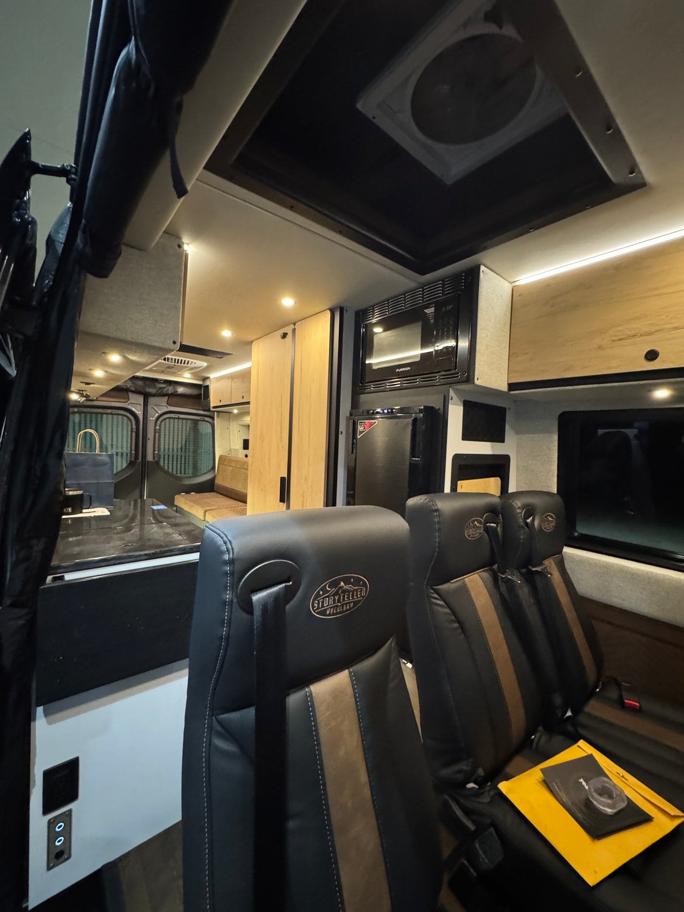 Modern camper van RV rental interior. Features comfortable black & brown seats, kitchen appliances, and overhead lighting for travel.
