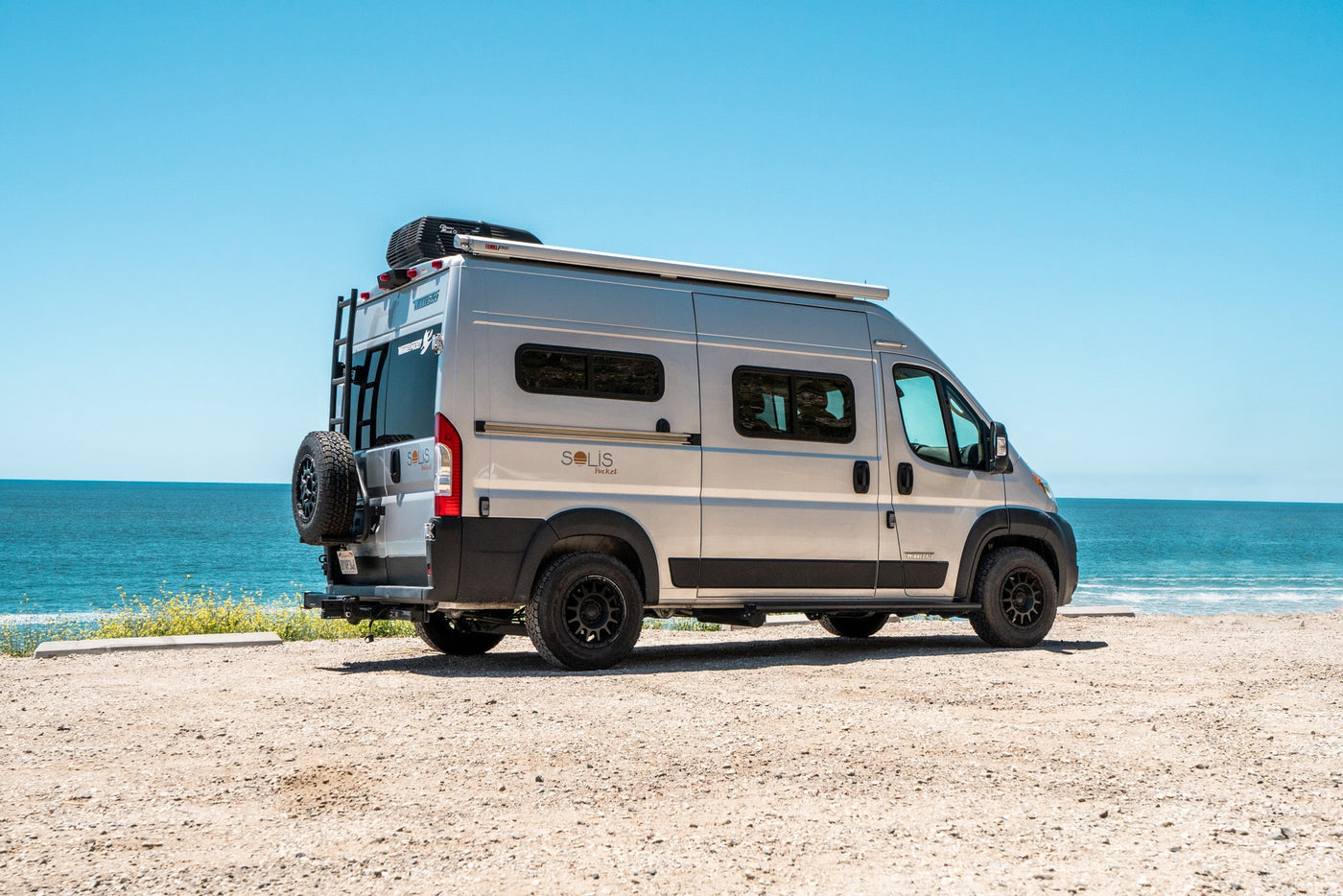 Silver Winnebago Solis camper van with roof AC and rear ladder, parked on a sunny dirt lot overlooking the blue ocean. Great for rv rental. — photo 6