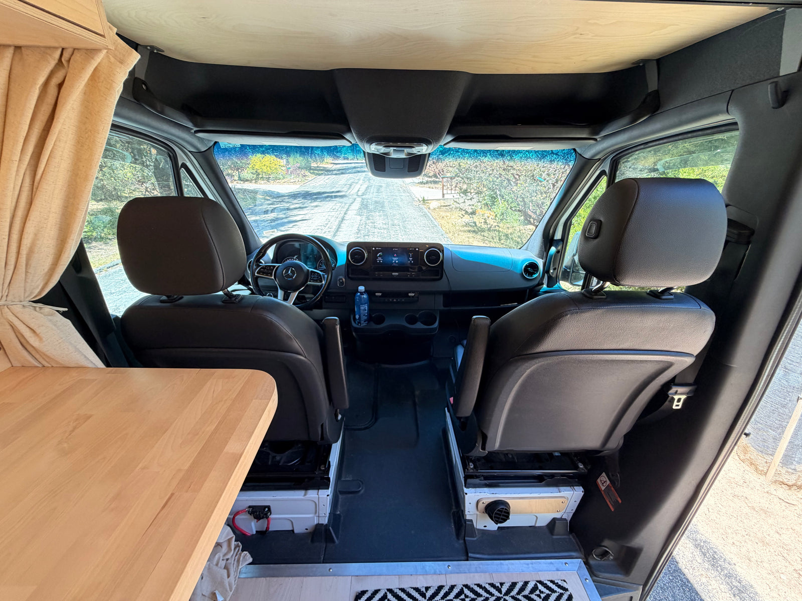 RV rental camper van interior. View from living area to front cockpit with dashboard, steering wheel, seats & wooden ceiling.