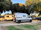 A silver Winnebago Revel 4x4 camper van at an RV park, surrounded by trees and other RVs. Perfect for your next rv rental adventure!