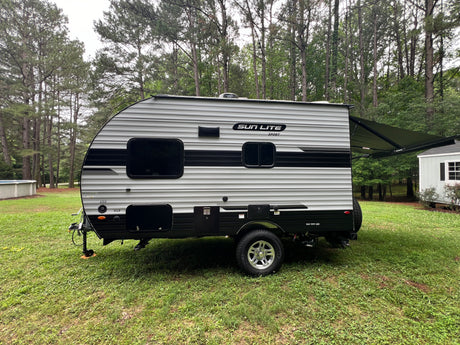 A grey and black striped Sun Lite Sport RV rental, a travel trailer with an extended awning, parked on green grass near trees.