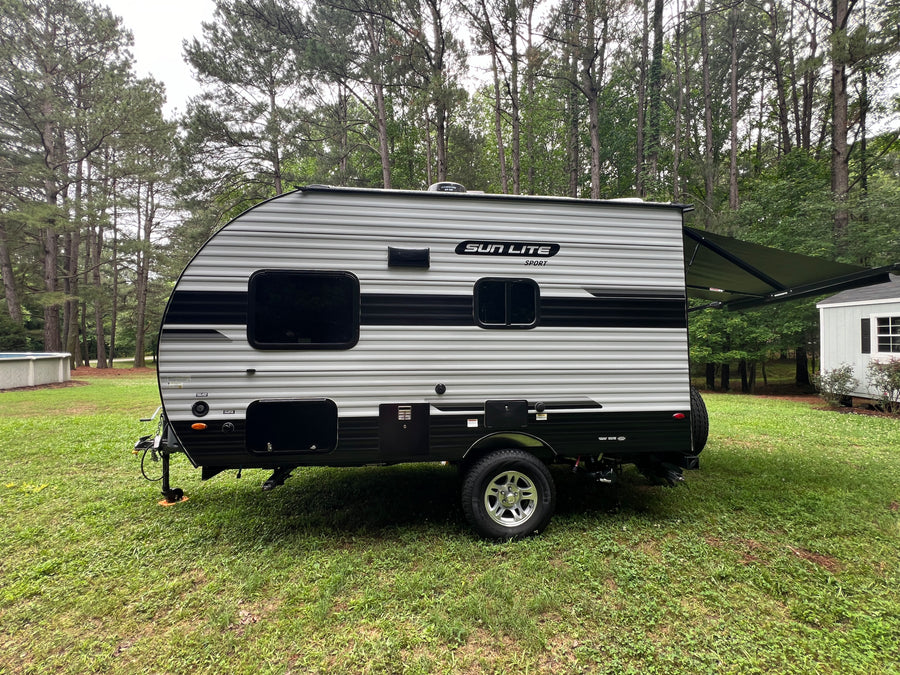 A grey and black striped Sun Lite Sport RV rental, a travel trailer with an extended awning, parked on green grass near trees.