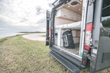This RV rental camper van's open rear shows a comfy bed & gear storage, set on a grassy beach shore.